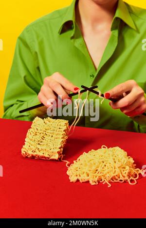 Image créative avec une femme triant des nouilles instantanées sur une table bleue sur une nappe rouge vif sur fond jaune. Photographie d'art pop. Banque D'Images