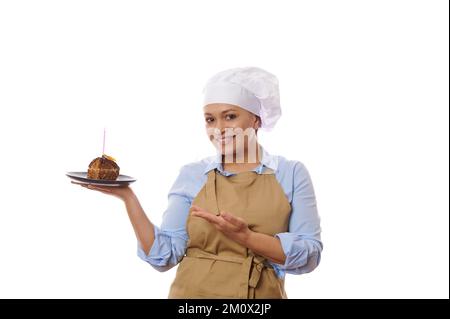 Charmante femme positive boulanger en tablier et chapeau, tient un morceau de délicieux gâteau au chocolat avec bougie, fond blanc Banque D'Images