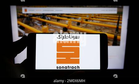 Personne tenant un téléphone portable avec le logo de la compagnie pétrolière et gazière algérienne Sonatrach sur l'écran en face de la page web d'affaires. Mise au point sur l'affichage du téléphone. Banque D'Images