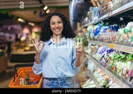 Un style de vie sain. Souriante jeune belle femme hispanique magasiner dans un supermarché, debout près des comptoirs, tenant le brocoli dans sa main et pointant vers l'appareil photo avec ses doigts ok. Banque D'Images
