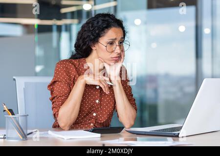 Épuisement professionnel. Jeune femme d'affaires latino-américaine assise fatiguée et contrariée à la table dans le bureau à l'ordinateur portable, beaucoup de travail, les délais. Banque D'Images