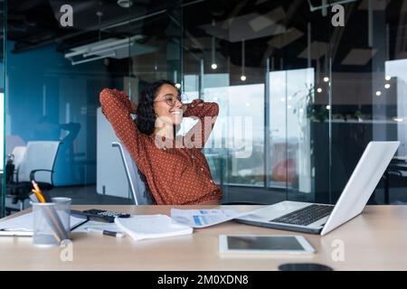 Une pause au travail. Résultat du travail réussi. Une jeune femme latino-américaine très belle est assise souriante dans le bureau au bureau, les mains derrière sa tête, reposant sur une chaise. Banque D'Images