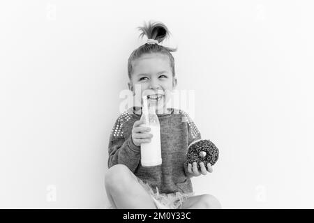 Jolie petite fille mange du beignet de chocolat avec une bouteille de lait. S'amuser, faire de la nourriture. Concept d'amoureux des bonbons. Photo en noir et blanc Banque D'Images
