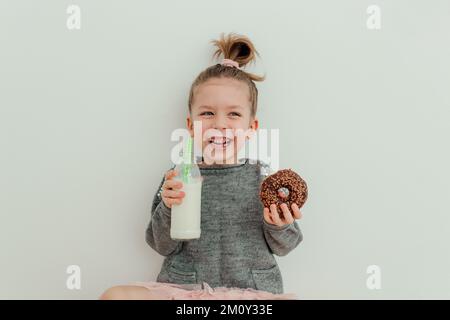 Jolie petite fille mange du beignet de chocolat avec une bouteille de lait. S'amuser, faire de la nourriture. Concept d'amoureux des bonbons. Style de vie Banque D'Images