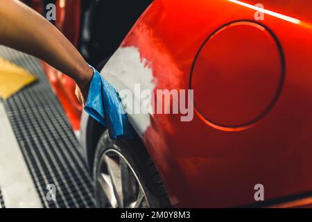 Main de mécanicien caucasien non reconnaissable à l'aide d'un chiffon bleu pour lisser la surface de la voiture rouge recouverte de peinture automobile avant le vernissage . Photo de haute qualité Banque D'Images