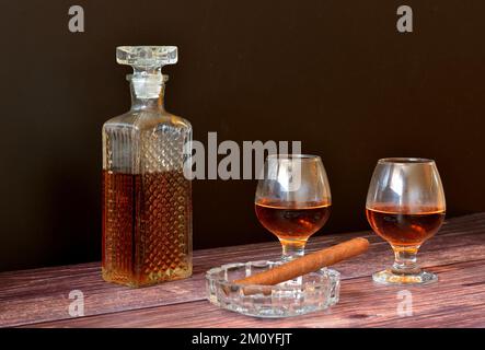 Cigare cubain dans un cendrier, deux verres et un carafe en cristal avec whisky sur une table en bois. Gros plan. Banque D'Images