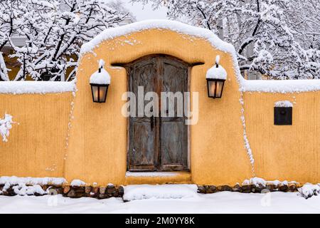 Hiver à Santa Fe, Nouveau-Mexique - mur en adobe recouvert de neige avec porte en bois rustique et lumières brillantes Banque D'Images