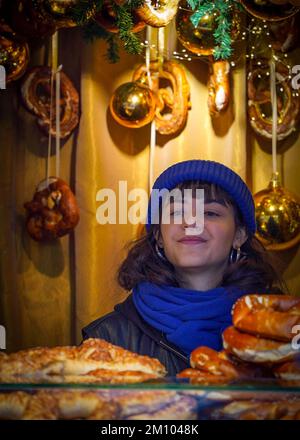 Une jeune fille vendeur de chapeau bleu et d'écharpe se tient à l'intérieur du kiosque illuminé de pâtisseries du marché de Noël encadré de décorations faites à la main du nouvel an Banque D'Images