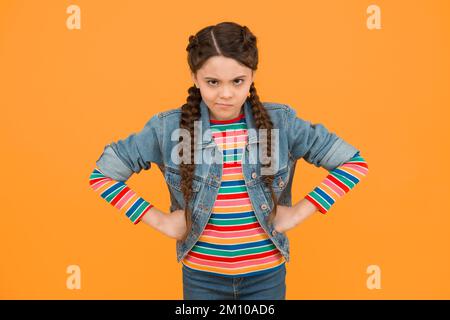 Dans la mauvaise humeur. Moody petit enfant fond jaune. Coiffure tressée pour bébé. Petite fille dans un style décontracté. Petit enfant avec les mains sur les hanches Banque D'Images