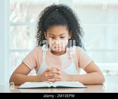 Guidage depuis un endroit plus haut. une petite fille ciblée priant en lisant une bible à la table à la maison. Banque D'Images