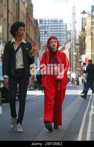 Cool Street style homme avec la coiffure afro, portant des jeans, veste et baskets blanches marche avec femme avec les cheveux rouges et la tracksuit rouge sur la rue de Londres Banque D'Images