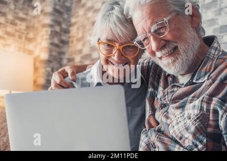 Couple mignon de personnes âgées assis sur le canapé en utilisant l'ordinateur portable ensemble faire du shopping et surfer sur le net. Deux personnes matures portant des lunettes dans le salon bénéficiant de la technologie. Portrait des aînés qui rient dans l'amour. Banque D'Images