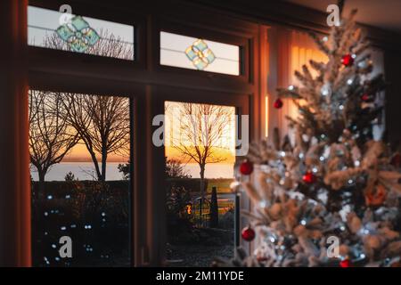 Des arbres hivernaux et une vue sur la mer pendant un coucher de soleil doré vu par une fenêtre de patio. Il y a un joli arbre de Noël dépoqué et givré avec Fair Banque D'Images