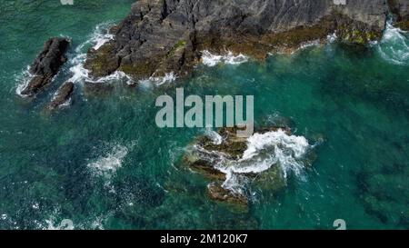 Rochers couverts de mousse noire au milieu des vagues turquoise de la mer celtique. Mousse de mer blanche sur les vagues. Eaux de l'océan Atlantique. Photo aérienne. Banque D'Images