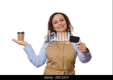 Jolie serveuse en tablier de chef beige, tient une boisson à emporter dans une tasse en papier et montre sur l'appareil photo une carte de crédit vierge Banque D'Images