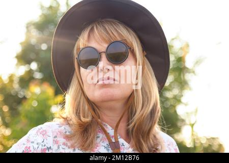 Réaliser un effet de flou sur le portrait extérieur d'une femme blonde d'âge moyen en chapeau et lunettes de soleil. Concept jeunesse, bien-être et style de vie. 40s femme sur fond de nature Banque D'Images