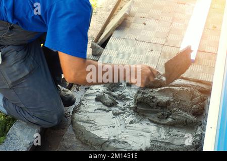 Le constructeur pose des carreaux autour du bâtiment. Briqueteuse professionnelle avec truelle au travail le jour ensoleillé de l'été. Flux de travail authentique... Banque D'Images