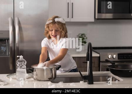 Petite gouvernante. L'enfant avec une éponge avec du liquide vaisselle fait la vaisselle à la cuisine maison en utilisant une éponge de lavage et le lave-vaisselle. Nettoyage Banque D'Images