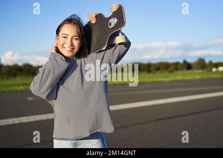 Portrait d'une jeune fille patineuse, debout à bord long et souriant à l'appareil photo. Femme asiatique avec planche à roulettes debout sur la route Banque D'Images