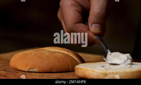 Préparation de délicieux hamburgers, gros plan d'un chef cuisinier qui met de la sauce mayonnaise blanche sur un pain hamburger. Concept gastronomique. Banque D'Images