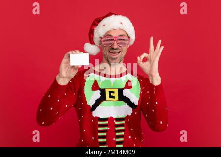 Santa man tient la carte de crédit. Studio portrait de type avec lunettes de fête porte chapeau de santa et chandail d'hiver. Homme avec chapeau de noël sur rouge isolé Banque D'Images