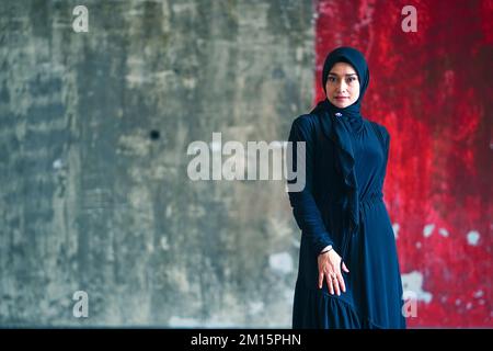 Belle femme indonésienne portant le hijab noir contre le mur rustique comme arrière-plan Banque D'Images