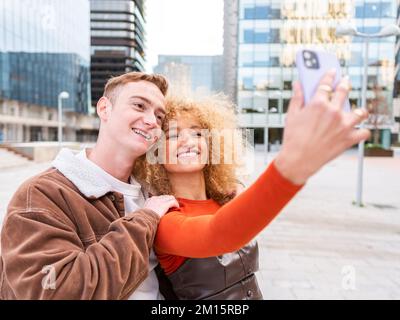 Un jeune couple multiracial ravie avec élégance et sourire tout en emportant un selfie sur un téléphone portable près des gratte-ciels contemporains Banque D'Images