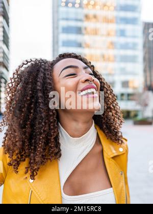 Jeune femme afro-américaine joyeuse et sûre d'elle-même avec de longs cheveux bouclés foncés dans une tenue élégante et souriante avec les yeux fermés pendant que vous visez Banque D'Images