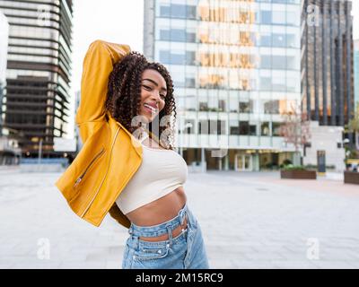 Jeune femme afro-américaine sûre d'elle-même avec de longs cheveux bouclés foncés dans une tenue élégante, souriant aux couleurs vives et regardant l'appareil photo tout en restant dans la chambre Banque D'Images