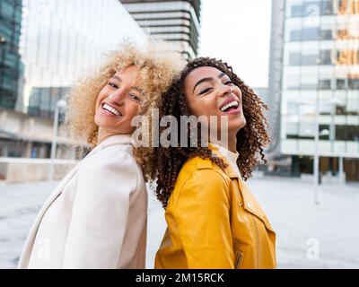 Vue latérale de jeunes filles gaies et stylées avec des cheveux bouclés souriant tout en se tenant dans la rue de la ville dos à dos et en regardant le chamer Banque D'Images