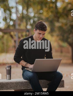 Étudiant masculin concentré dans une tenue décontractée assis sur un banc de pierre près de thermomug tout en travaillant à distance via un ordinateur portable dans le parc d'automne Banque D'Images