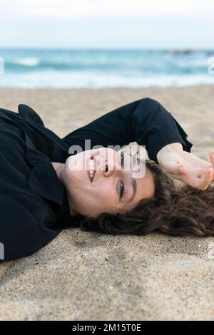 Vue latérale d'une jeune femme heureuse avec des cheveux bouclés souriant et couché sur une plage de sable Banque D'Images