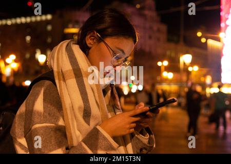 Vue latérale de la jeune femme hispanique en extérieur et lunettes de texte sur un téléphone portable tout en se tenant dans la rue urbaine la nuit en hiver Banque D'Images