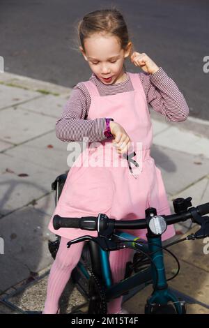 Bonne fille en robe rose assis sur le vélo et vérifier l'heure dans la rue de ville en journée Banque D'Images