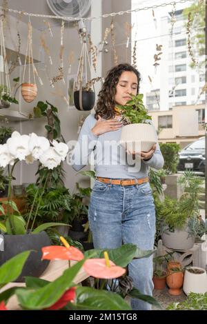 Positive jeune hispanique femelle acheteur dans les vêtements décontractés debout dans la boutique florale près de la fenêtre tout en choisissant la plante en pot Banque D'Images