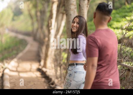 Jeune couple multiracial positif en vêtements décontractés tenant les mains et marchant sur la passerelle dans le parc Banque D'Images