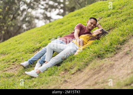Joyeux jeune homme et femme dans des vêtements décontractés souriant et regardant les uns les autres tout en se relaxant sur l'herbe dans le parc pendant la date romantique Banque D'Images