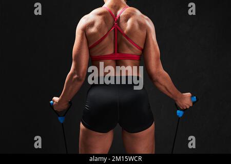 Vue arrière d'une courte femme musclée non reconnaissable, dans un vêtement de sport serré, s'entraîner avec un câble d'entraînement tout en restant debout sur fond noir Banque D'Images