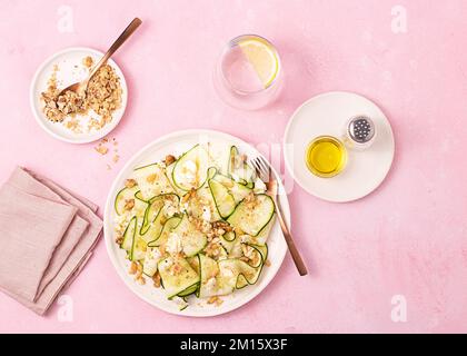 De dessus salade fraîche de courgettes fines tranchées mûres, fromage feta et noix de pin dans une assiette blanche sur fond rose de table Banque D'Images