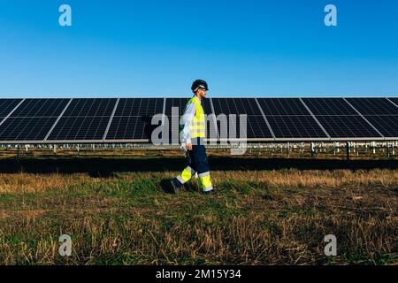 Vue latérale de l'installateur dans un casque de protection et vêtements de travail marchant sur l'herbe près des panneaux solaires sous un ciel bleu sans nuages Banque D'Images