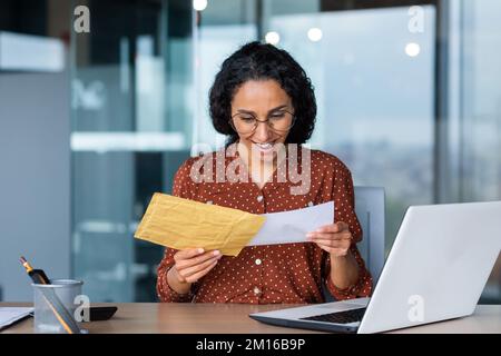 Une jeune femme latino-américaine heureuse a reçu une facture avec un salaire gagné, une récompense en espèces, de bonnes nouvelles. Il est assis au bureau, en tenant une enveloppe avec une lettre entre ses mains. Banque D'Images