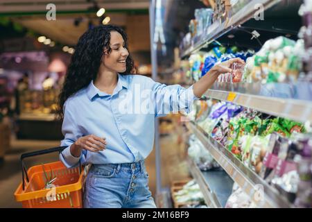 Une jeune belle femme hispanique achète des tomates cerises dans un supermarché. Elle se tient souriante près du réfrigérateur, le département des légumes avec un panier pour les provisions. Banque D'Images