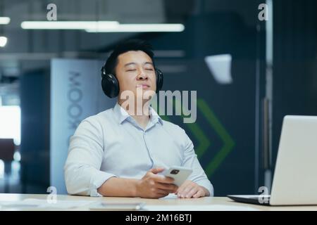 Repos, pause au travail. Un jeune homme asiatique est assis au bureau, avec un casque, et écoute de la musique à partir d'un téléphone portable. Il ferme les yeux, détendu. Banque D'Images