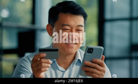 Happy Asian homme client acheteur homme d'affaires utiliser l'application bancaire mobile téléphone au bureau tenir carte de crédit en plastique entrer code sécurisé acheter des marchandises dans la boutique Internet Banque D'Images