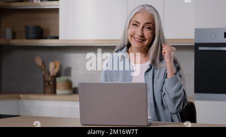 Grand-mère d'âge moyen heureux s'asseoir à la maison utiliser ordinateur portable vérifier e-mail lire les nouvelles positives obtenir la vente offre sentir l'excitation faire oui gestuelle céléra Banque D'Images