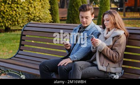 Couple caucasien deux jeunes partenaires assis sur le banc utiliser téléphone cellulaire gadgets faire défiler les pages Web de vérifier les réseaux sociaux montrent des images les uns les autres Banque D'Images