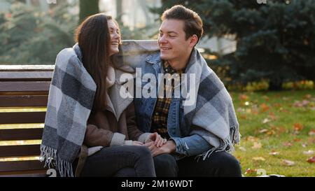 Mignon couple aimant s'asseoir sur le banc chauffant l'un l'autre couvrant couverture de laine dehors dans froid gelé temps automnal se sentir soin amour embrassant drôle parler Banque D'Images