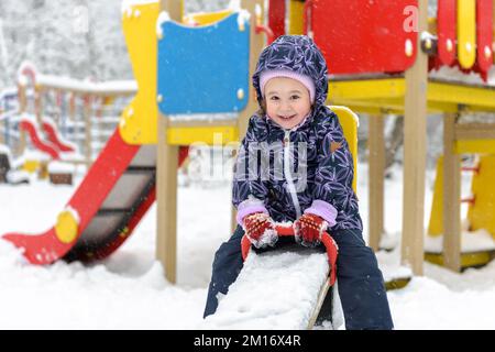 Enfant sur la mer en hiver, petit enfant s'amusant sur une aire de jeux enneigée. Bébé fille joue et regarde la caméra dans le parc urbain. Thème neige, jeu, Christma Banque D'Images