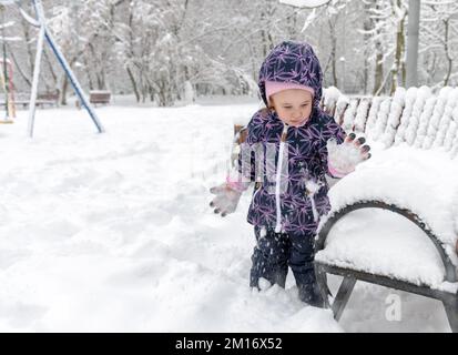 Les enfants jouent avec la neige en hiver, les petits enfants se promène dans un parc urbain enneigé pendant les chutes de neige. Bébé fille est sur fond de neige. Thème du froid, du gel, de l'hiver, Banque D'Images