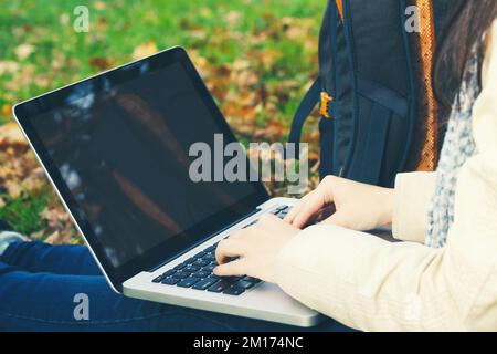 une fille utilise un ordinateur portable tout en étant assise sur l'herbe dans le parc d'automne. Vide pour le design sur l'écran de l'ordinateur portable. Banque D'Images